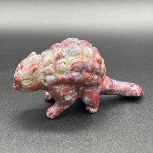 Könnte beinhalten: Eine geschnitzte Dinosaurierfigur aus Stein in Rot-, Weiß- und Beigetönen. Die Skulptur hat einen strukturierten Rücken, kleine Beine und einen langen Schwanz. Der Dinosaurier steht auf einer dunklen Oberfläche.