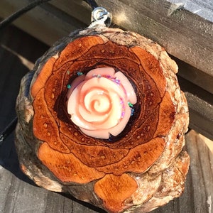 May include: A wooden pendant with a pink rose resin inlay. The wood is a dark brown with a natural wood grain pattern. The pendant is attached to a black cord.