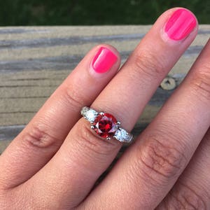 May include: A silver ring with a large red gemstone surrounded by two smaller white gemstones.