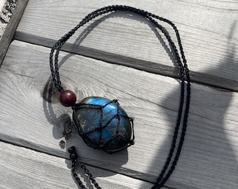 May include: A black cord necklace with a dark blue labradorite stone wrapped in black macrame. The stone has a iridescent sheen.