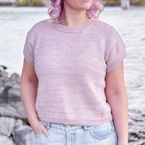 May include: A light pink knit top with short sleeves. The top has a crew neck and a slightly textured appearance. The person is wearing light wash denim jeans. The background includes a river, rocks, and a bridge.