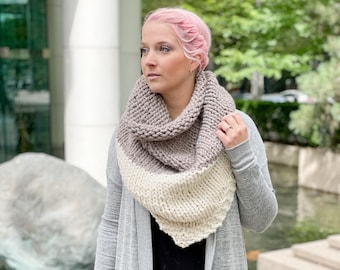 May include: A woman wearing a gray cardigan, a black shirt, and light blue jeans. She is wearing a gray, white, and beige knitted infinity scarf.