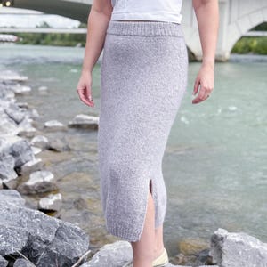 May include: A light gray knitted skirt with a side slit, paired with a white tank top and light yellow sneakers. The skirt is knee-length and appears to be made of a soft, textured yarn. The background features a river and a bridge.