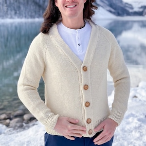 May include: A cream-colored knitted cardigan with a V-neck and wooden buttons. The cardigan is worn by a person standing in front of a snowy mountain landscape.