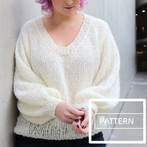 May include: A white knitted sweater with a v-neck and long sleeves. The sweater has a loose fit and a slightly textured knit. The model is wearing the sweater with black pants. The word "PATTERN" is visible in the lower right corner of the image.