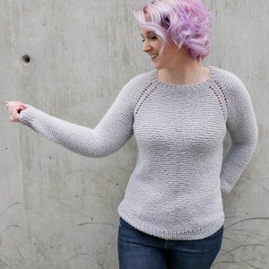 May include: A gray knitted sweater with a round neckline and long sleeves. The sweater has a subtle texture and a delicate lace pattern around the neckline and shoulders. The model is wearing blue jeans.