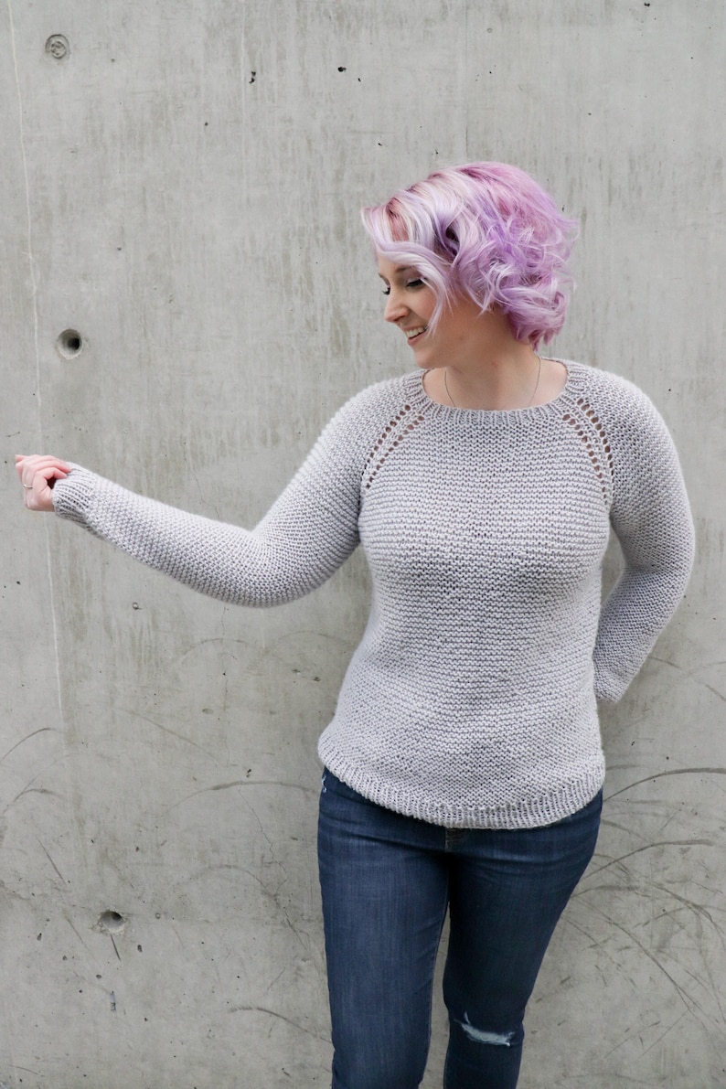 May include: A gray knitted sweater with a round neckline and long sleeves. The sweater has a subtle texture and a delicate lace pattern around the neckline and shoulders. The model is wearing blue jeans.