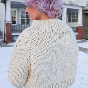 May include: A close-up of a white knitted sweater with a high neck and long sleeves. The sweater is cropped and has a relaxed fit.