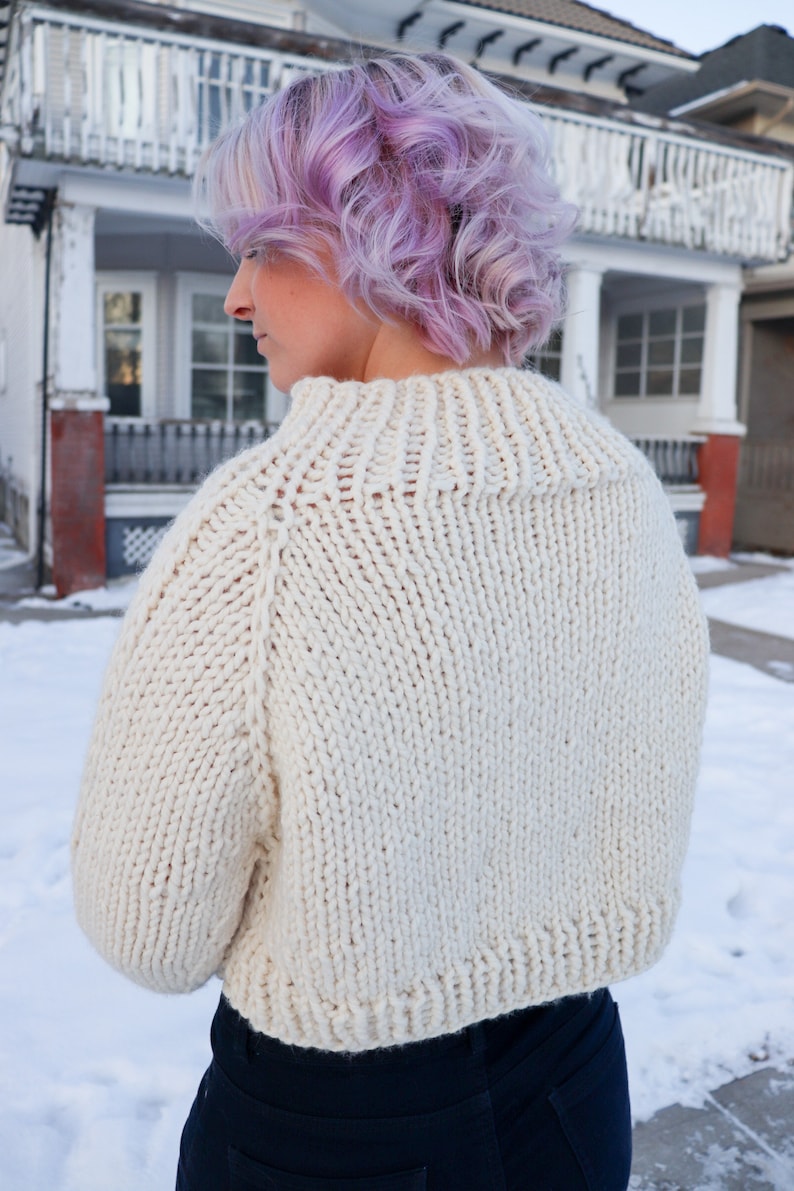 May include: A close-up of a white knitted sweater with a high neck and long sleeves. The sweater is cropped and has a relaxed fit.