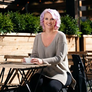May include: A woman with pink hair sits at a cafe table, wearing a grey sweater and black pants. She holds a white coffee cup in her hands.