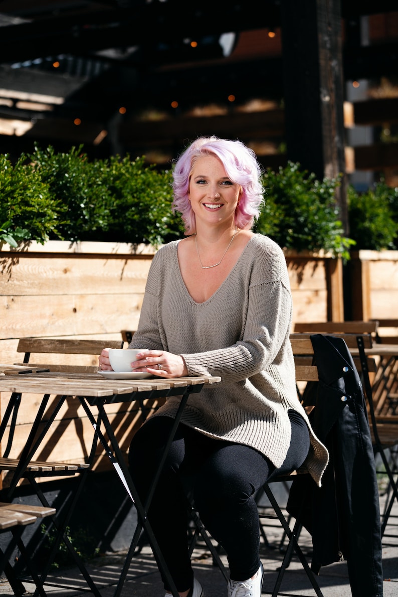 May include: A woman with pink hair sits at a cafe table, wearing a grey sweater and black pants. She holds a white coffee cup in her hands.