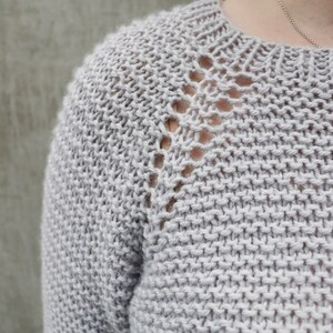 May include: A close-up of a light gray knitted sweater with a delicate openwork pattern around the neckline.