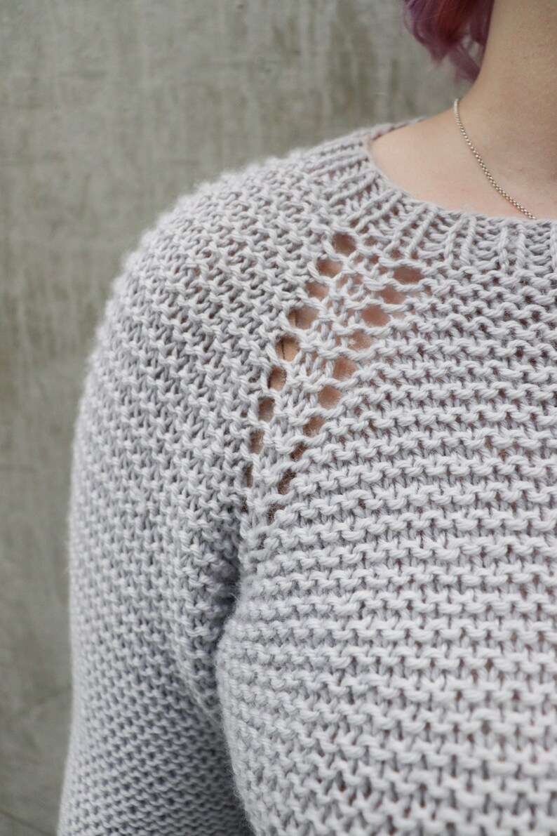 May include: A close-up of a light gray knitted sweater with a delicate openwork pattern around the neckline.