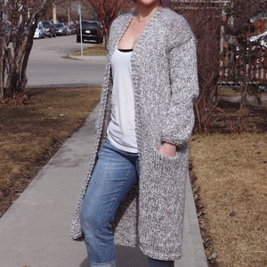 May include: A woman with purple hair is wearing a long, grey and white knit cardigan, blue jeans, and white trainers. She is standing on a pavement.