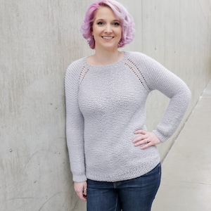 May include: A woman with short, pink hair is wearing a light gray sweater with a round neckline and long sleeves. She is also wearing dark blue jeans.