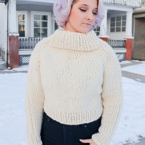 May include: A white knitted turtleneck sweater with long sleeves. The sweater is cropped and has a relaxed fit.