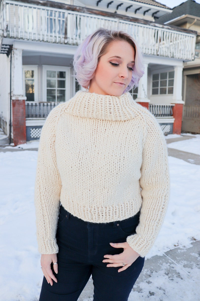 May include: A white knitted turtleneck sweater with long sleeves. The sweater is cropped and has a relaxed fit.