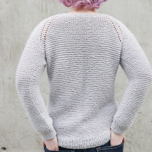 May include: A gray knitted sweater with a round neckline and long sleeves. The sweater has a subtle texture and is worn by a person with pink hair.