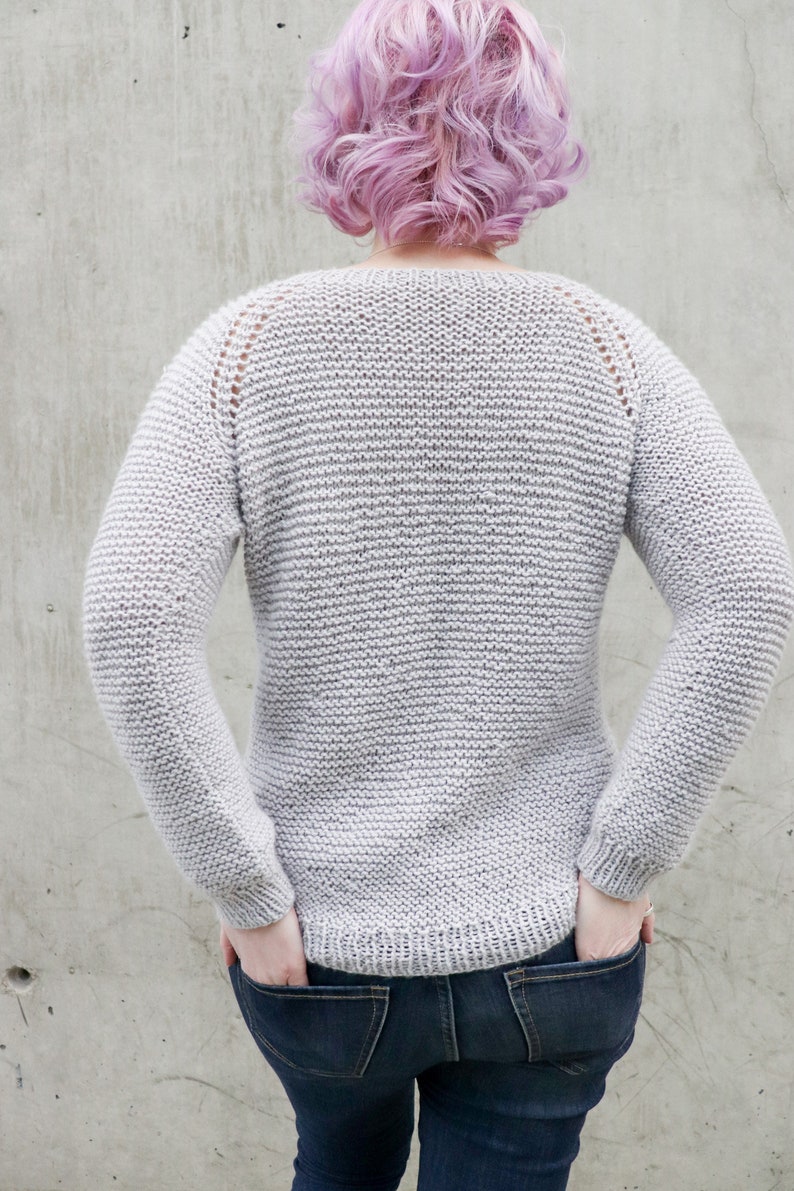 May include: A gray knitted sweater with a round neckline and long sleeves. The sweater has a subtle texture and is worn by a person with pink hair.