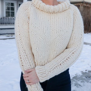May include: A white knitted sweater with a turtleneck and long sleeves. The sweater is cropped and has a relaxed fit.
