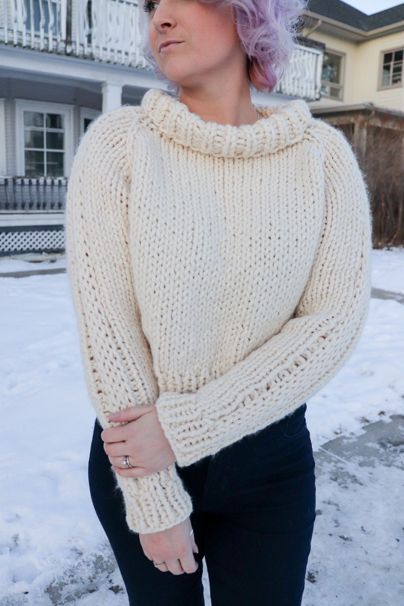 May include: A white knitted sweater with a turtleneck and long sleeves. The sweater is cropped and has a relaxed fit.