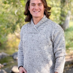 May include: A light gray knit sweater with a shawl collar. The sweater has a speckled pattern and long sleeves. The person is wearing a black watch and a black bracelet. The background is a blurred outdoor scene.