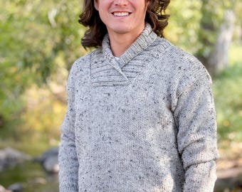 May include: A light gray knit sweater with a shawl collar. The sweater has a speckled pattern and long sleeves. The person is wearing a black watch and a black bracelet. The background is a blurred outdoor scene.