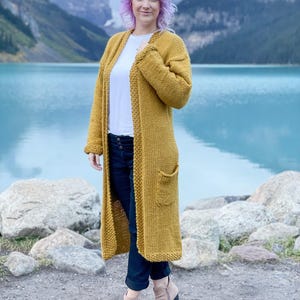 May include: A long, mustard yellow cardigan with a loose fit and pockets. The cardigan is knitted and has a simple design.