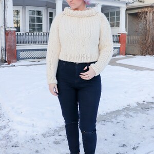 May include: A white cropped sweater with a high neck and a loose fit. The sweater is worn with dark blue jeans and brown ankle boots.