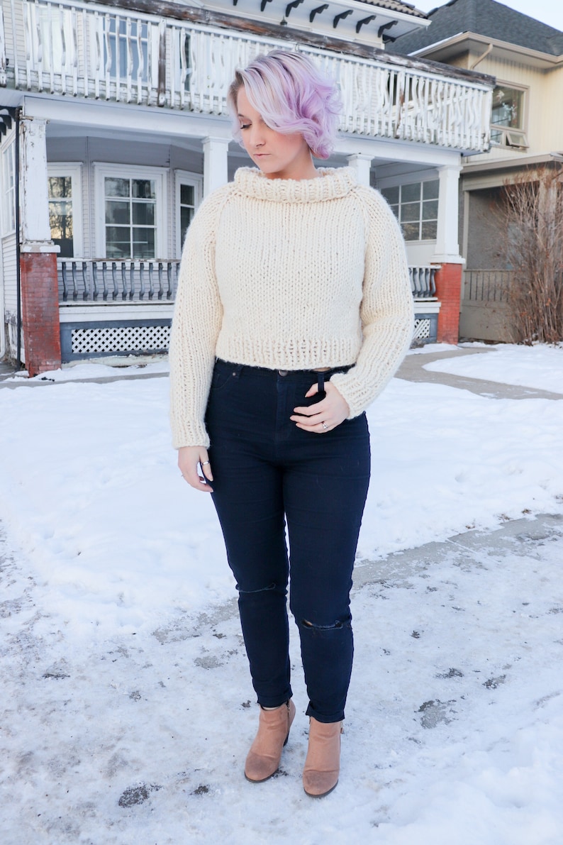 May include: A white cropped sweater with a high neck and a loose fit. The sweater is worn with dark blue jeans and brown ankle boots.