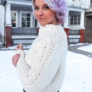 May include: A woman with short, lavender hair is wearing a white, chunky knit sweater with a high neck. She is standing in front of a house with snow on the ground.