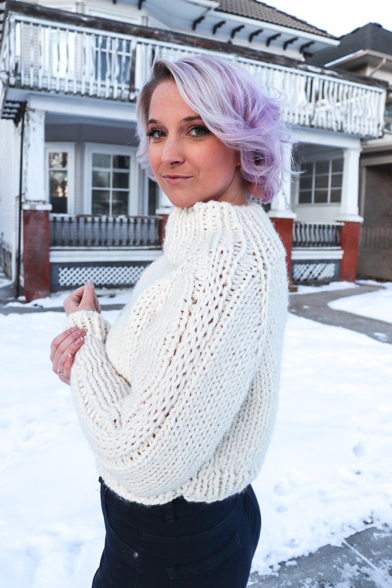 May include: A woman with short, lavender hair is wearing a white, chunky knit sweater with a high neck. She is standing in front of a house with snow on the ground.