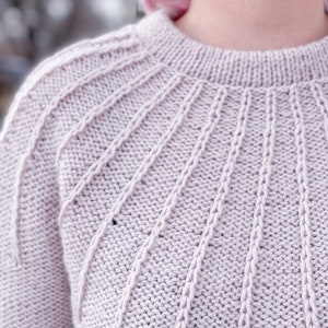 May include: Close-up of a light pink knitted jumper with a detailed cable knit pattern around the neckline.