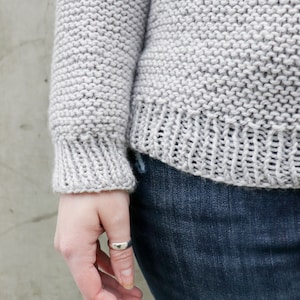 May include: A close-up of a gray knitted sweater with a ribbed cuff. The sweater is worn over a pair of blue jeans.