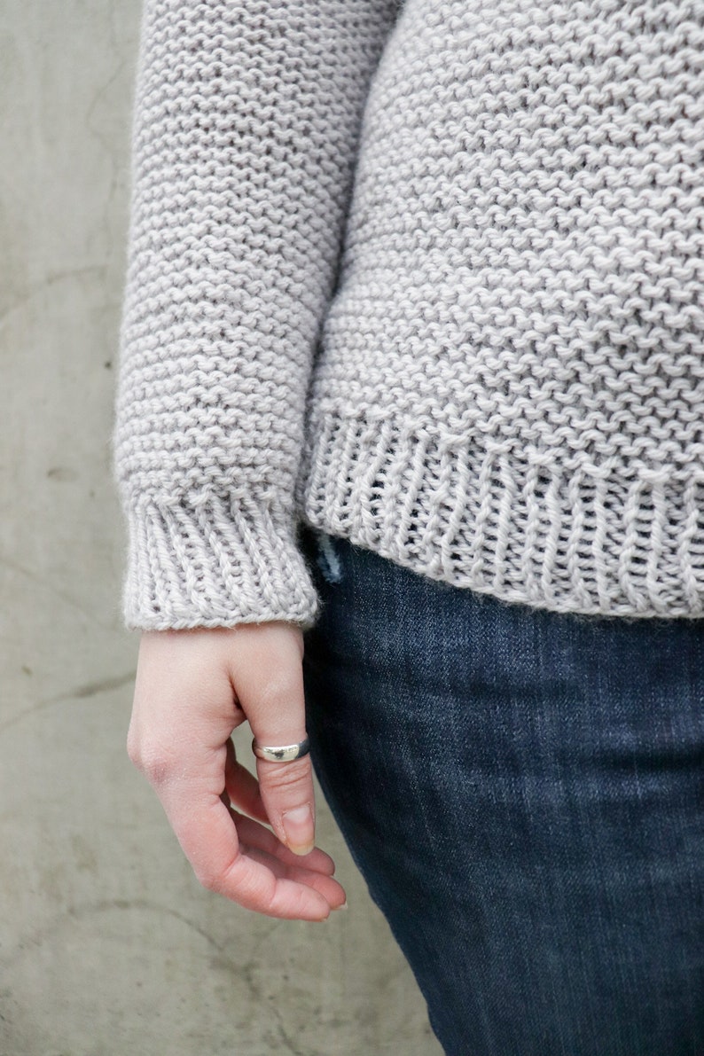 May include: A close-up of a gray knitted sweater with a ribbed cuff. The sweater is worn over a pair of blue jeans.