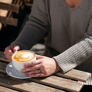 May include: A white ceramic mug with a latte art design, held by a person wearing a gray sweater, sitting at a wooden table.