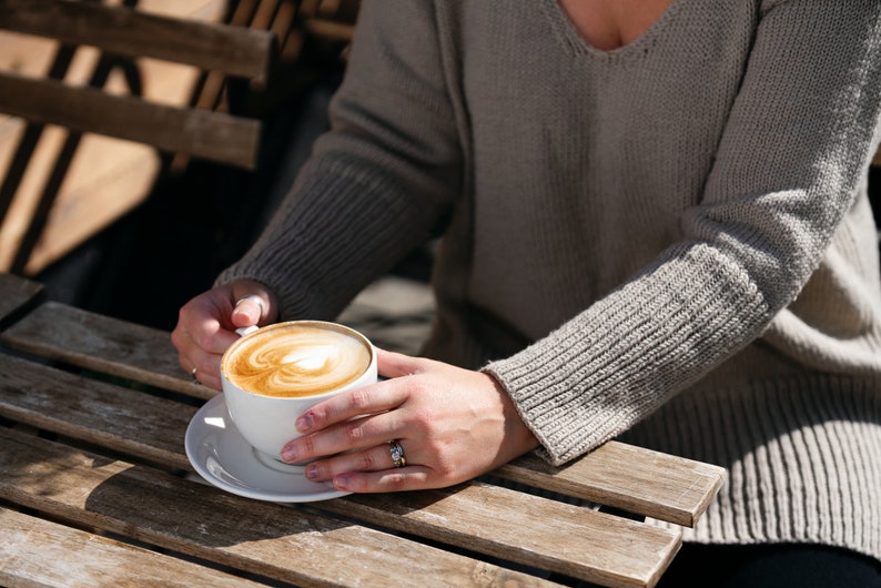 May include: A white ceramic mug with a latte art design, held by a person wearing a gray sweater, sitting at a wooden table.