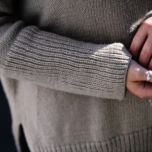 May include: A close-up of a person's hands wearing two silver rings. The person is wearing a light brown knitted sweater.