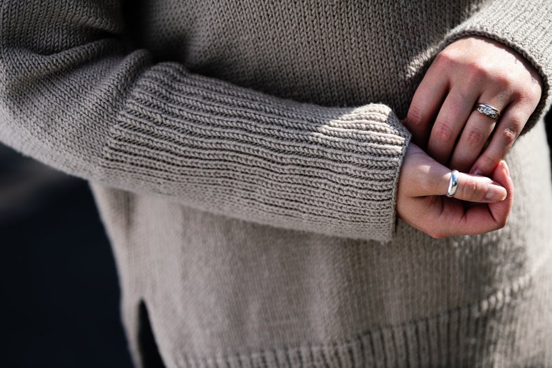 May include: A close-up of a person's hands wearing two silver rings. The person is wearing a light brown knitted sweater.