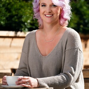 May include: A woman with pink hair is sitting at a table outside. She is wearing a gray sweater and a silver necklace. She is holding a white coffee cup in her hands.