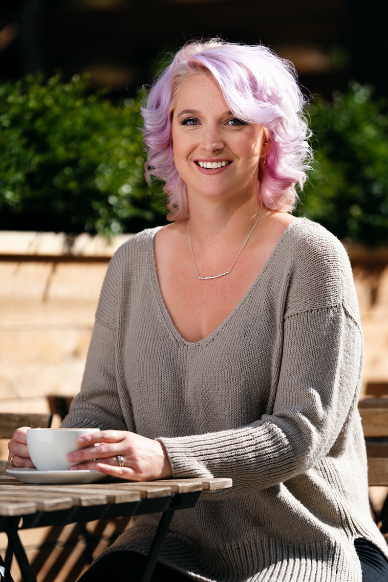 May include: A woman with pink hair is sitting at a table outside. She is wearing a gray sweater and a silver necklace. She is holding a white coffee cup in her hands.