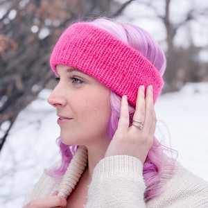 Op de afbeelding: Een roze gebreid hoofdband met een zachte, pluche textuur. De hoofdband wordt gedragen door een persoon met paars haar.