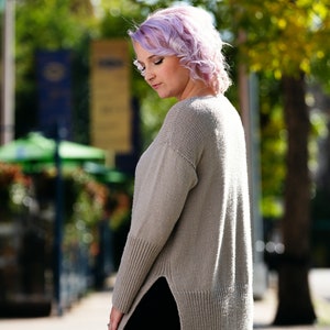 May include: A woman with short, curly, lavender hair is wearing a long, light brown, knitted sweater with a side slit. She is wearing black pants and is standing on a sidewalk.