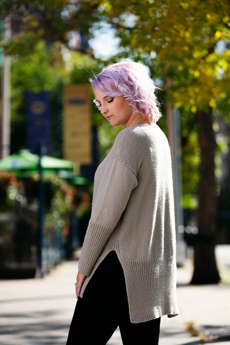 May include: A woman with short, curly, lavender hair is wearing a long, light brown, knitted sweater with a side slit. She is wearing black pants and is standing on a sidewalk.