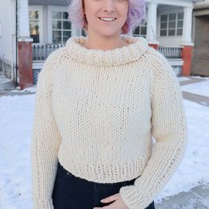 May include: A white knitted sweater with a turtleneck and long sleeves. The sweater is cropped and has a loose fit.