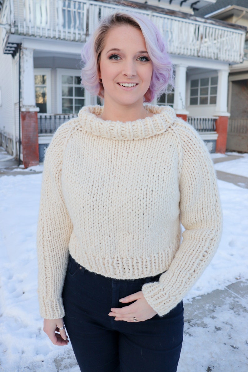 May include: A white knitted sweater with a turtleneck and long sleeves. The sweater is cropped and has a loose fit.