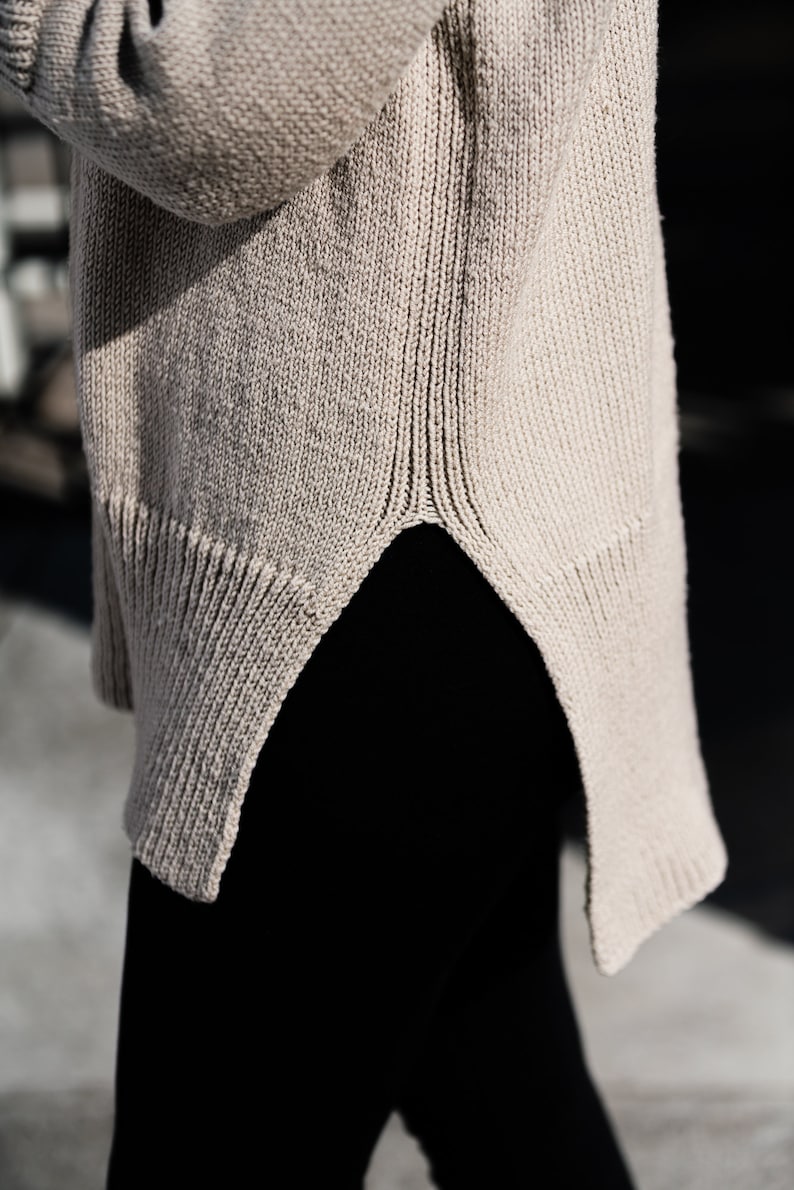 May include: A beige knitted sweater with a high neckline and a long, slightly asymmetrical hem.
