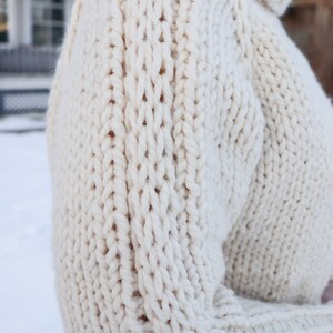 May include: A close-up of a cream-colored knitted sweater with a chunky cable knit pattern.