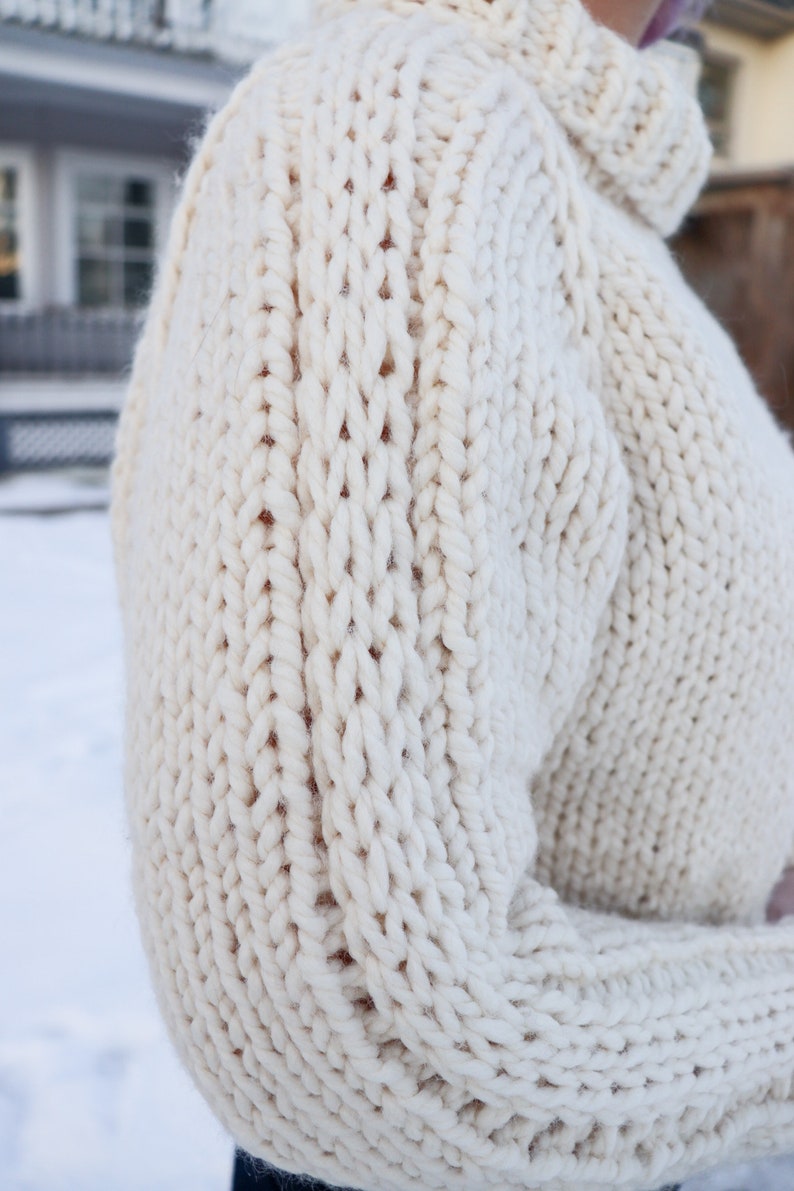May include: A close-up of a cream-colored knitted sweater with a chunky cable knit pattern.