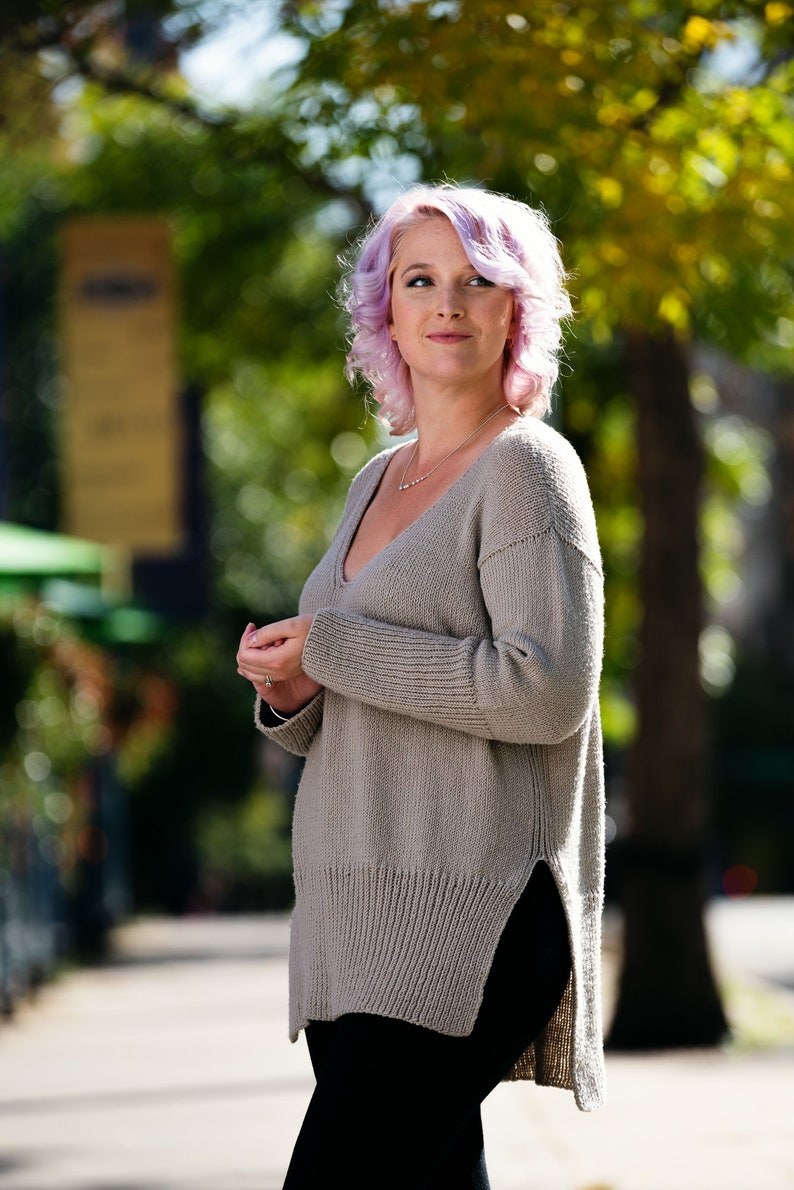 May include: A woman with pink hair is wearing a light gray, long-sleeved sweater with a v-neck and a side slit. She is wearing black pants.